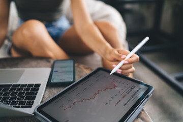 Close-up of female hand with stylus pointing at rising crypto graph on tablet, multitasking with...
