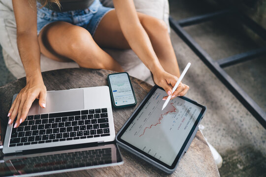 Woman sitting cross-legged, using stylus on tablet to analyze crypto charts, with smartphone and laptop nearby, working remotely in relaxed digital workspace