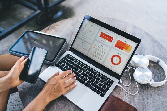 Close-up of female freelancer analyzing colorful business charts on laptop screen while holding smartphone, with tablet and headphones on desk - Powered by Adobe