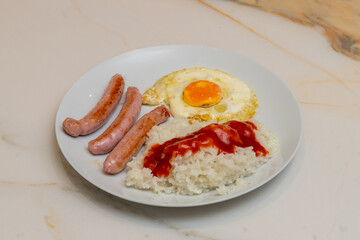 A combination plate with three sausages, white rice with tomato and a fried egg on a veined white marble table