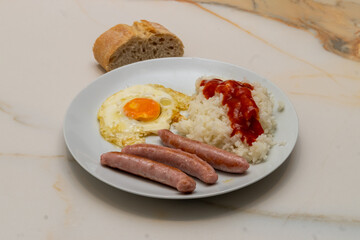 A combination plate with three sausages, white rice with tomato and a fried egg with a piece of bread on a veined white marble table