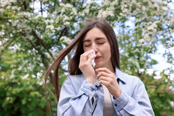 Woman with paper tissue suffering from seasonal pollen allergy near blossoming tree on spring day,...