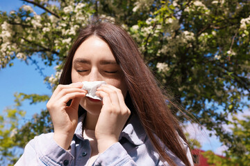 Woman with paper tissue suffering from seasonal pollen allergy near blossoming tree on spring day
