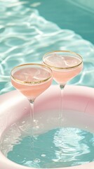 Two pink cocktails in coupe glasses with a gold rim, floating on an inflatable pastel swim ring in a pool with light blue water.