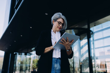 Confident older businesswoman in smart casual outfit focused on her tablet near office building,...