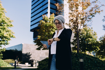 Mature businesswoman with tablet looking at wristwatch in city park, representing time management,...