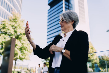 Senior woman entrepreneur using smartphone for video call in urban environment, expressing connection, digital professionalism and modern remote communication tools