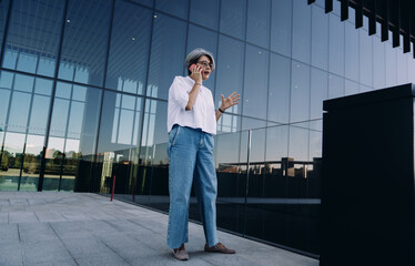 Confident senior woman gesturing during phone conversation outside modern office. Expressive communication, strong presence and dynamic body language in corporate environment