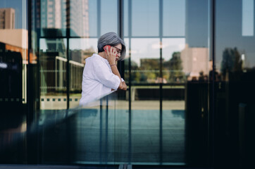 Thoughtful senior businesswoman leaning on railing during phone conversation. Reflective mood, calm...