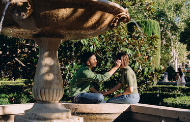 Man gently places flower in woman’s hair while sitting by the fountain, romantic gesture in lush...