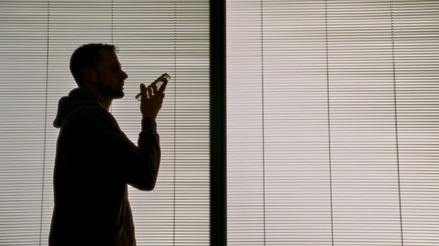 Silhouette of man recording voice message on smartphone standing next to window. Office or apartment window with venetian blinds. Young male holding phone to mouth and speaks sending sound sms