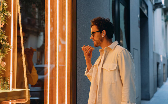 Caucasian Handsome Man with wireless earphones and smartphone app using voice command near store window. Smart assistant, hands-free interaction, and modern digital lifestyle concept