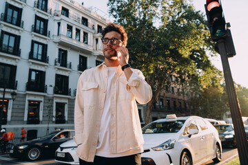 young male entrepreneur in glasses and light jacket takes an important phone call in a dynamic city setting. White taxis and modern buildings create a vibrant, fast-paced business atmosphere
