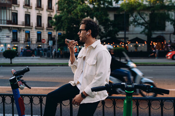 stylish handsome man in glasses and a beige jacket sits by the street, sending a voice message on...