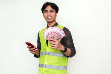 Smiling Asian construction worker holding cash and using smartphone, isolated on white background....