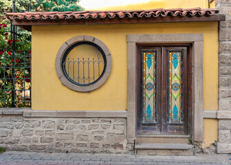 Facade of a traditional house at an old city. Yellow wall with a stone base, stained glass door, and a round window with an iron grille