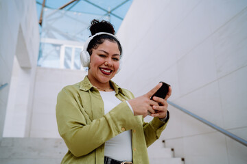 Cheerful woman enjoys seamless technology interaction, using her smartphone while listening to music on wireless headphones. She stays connected,browsing online content and engaging with digital media