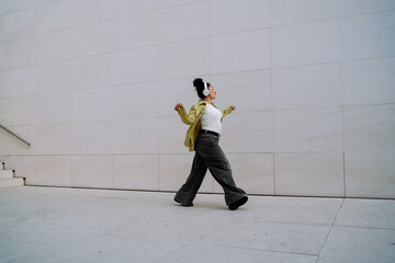 Young woman in wireless headphones dances freely in minimalist urban setting. Femal tech user...