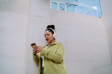 Confident young Latina woman listens to music with wireless headphones while checking her phone....