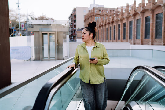 thoughtful latina woman on an escalator checks her banking app on a smartphone. She reviews transactions, manages finances, and prepares for online purchases while commuting in an urban setting