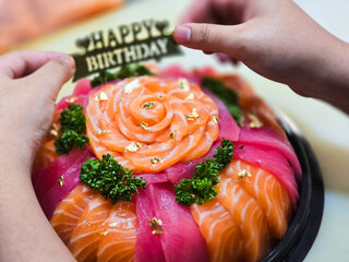 Birthday sashimi platter decorated with fresh salmon and tuna slices, arranged in a rose pattern, gold leaf flakes, parsley, and a “Happy Birthday” sign being placed by hand.