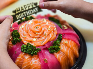 Birthday sashimi platter decorated with fresh salmon and tuna slices, arranged in a rose pattern, gold leaf flakes, parsley, and a “Happy Birthday” sign being placed by hand.