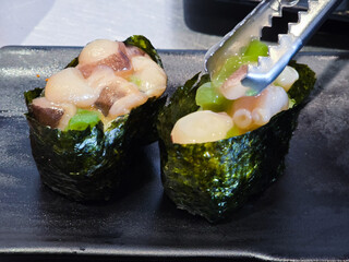 Gunkan sushi with fresh raw octopus, seaweed wrap, and green garnish being served with metal tongs on a black plate in a Japanese restaurant.