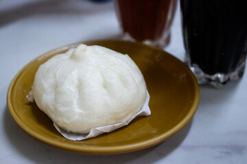 A freshly steamed Chinese pork bun served on a ceramic plate, with iced beverages in the background on a marble table.