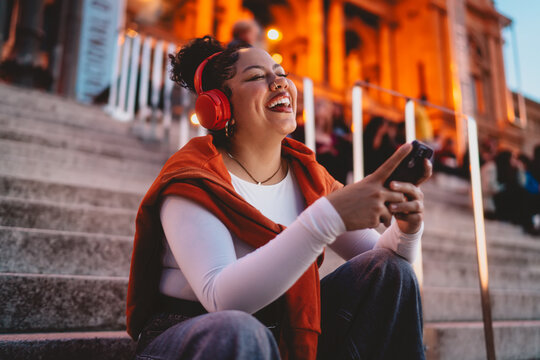joyful hispanic woman wearing wireless headphones laughs while engaging with her smartphone. The city lights illuminate the urban scene, enhancing the digital experience of music and social media