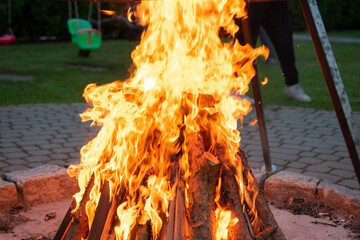 Fire is burning in a pile of wood, with a person standing behind it. The fire is very large and is...