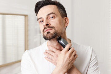 Handsome man shaving with trimmer at home