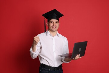 Happy student with laptop after graduation on red background