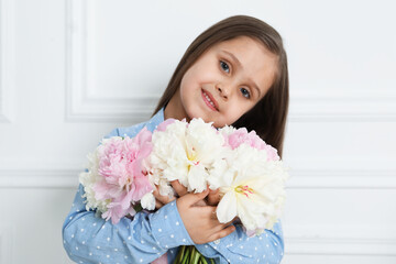 Cute little girl with bouquet of beautiful peonies near white wall indoors