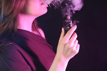 Fototapeta premium Woman using electronic smoking device in neon lights on black background, closeup