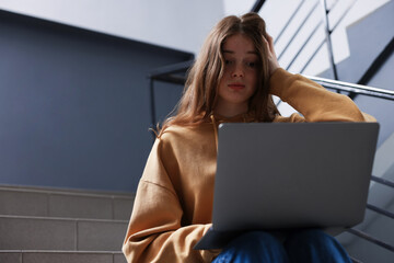 Cyber bullying. Upset teenage girl with laptop on stairs indoors. Space for text