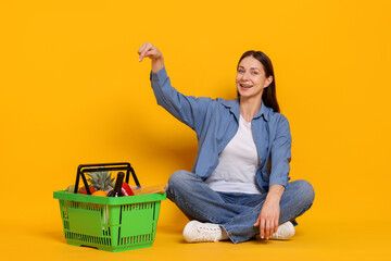 Smiling woman pointing at shopping basket full of products on yellow background