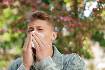 Young man with paper tissue suffering from seasonal pollen allergy near blossoming tree on spring...
