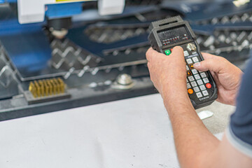 Person operating a handheld industrial controller, likely for machinery or equipment, in an indoor workspace with heavy machinery and artificial lighting