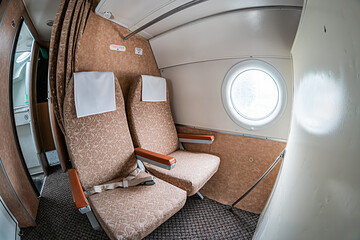 Small aircraft cabin interior from front seat, featuring at least three light-colored seats possibly cream or light brown with headrests Walls have fabric coverings and an oval window is visible o