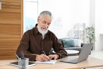 Senior man taking notes while learning online at desk indoors. Self-study