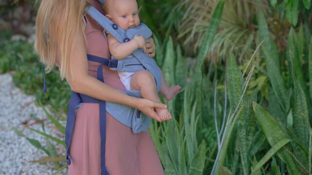 Young mother walking in a green park with her baby son sitting face forward in an ergonomic baby carrier, enjoying a sunny day together outdoors. Slowmotion video.