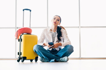 Bored stylish woman with yellow suitcase seated at airport waiting area using smartphone during...