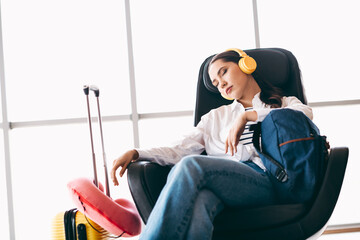 Young female traveler resting at airport lounge with headphones and luggage, embracing comfort...
