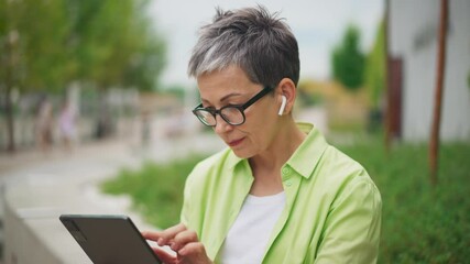 Elderly woman with short gray hair and glasses adjusting an earbud while focusing on a tablet outdoors, with greenery and walkway in the background. Caucasian female wearing wireless headphone.