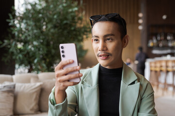 Portrait of nonbinary genderfluid young Asian gay businessman using phone in cafe