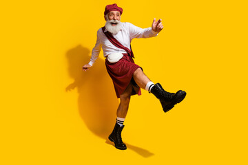 Smiling elder man in Scottish attire making joyful gestures against vibrant yellow studio backdrop