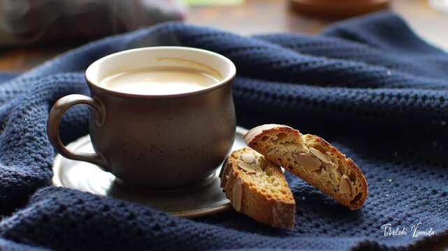A cup of coffee and biscotti on a saucer with a blue knitted blanket in the background still life