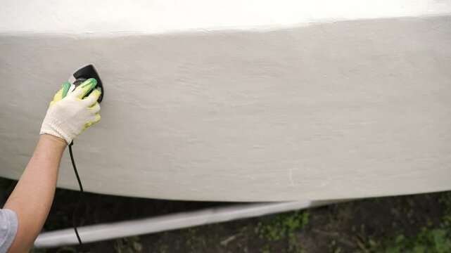 Professional worker carefully polishing white boat hull using orbital sander, working meticulously to restore smooth surface and maintain nautical vessel's exterior