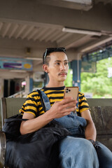 Portrait of nonbinary genderfluid young Asian gay tourist man in bus terminal using phone