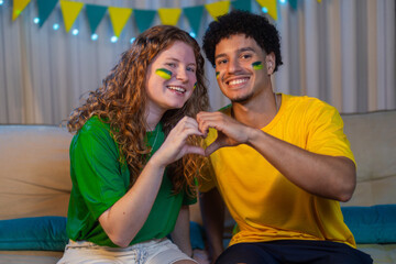 Multiracial couple sitting on sofa with face paint, smiling and forming a heart shape with hands during a Brazilian World Cup game.
